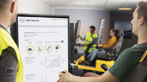 A guy is practicing in a simulator with a teacher standing next to him. In the background girl in simultaneous with teacher next to